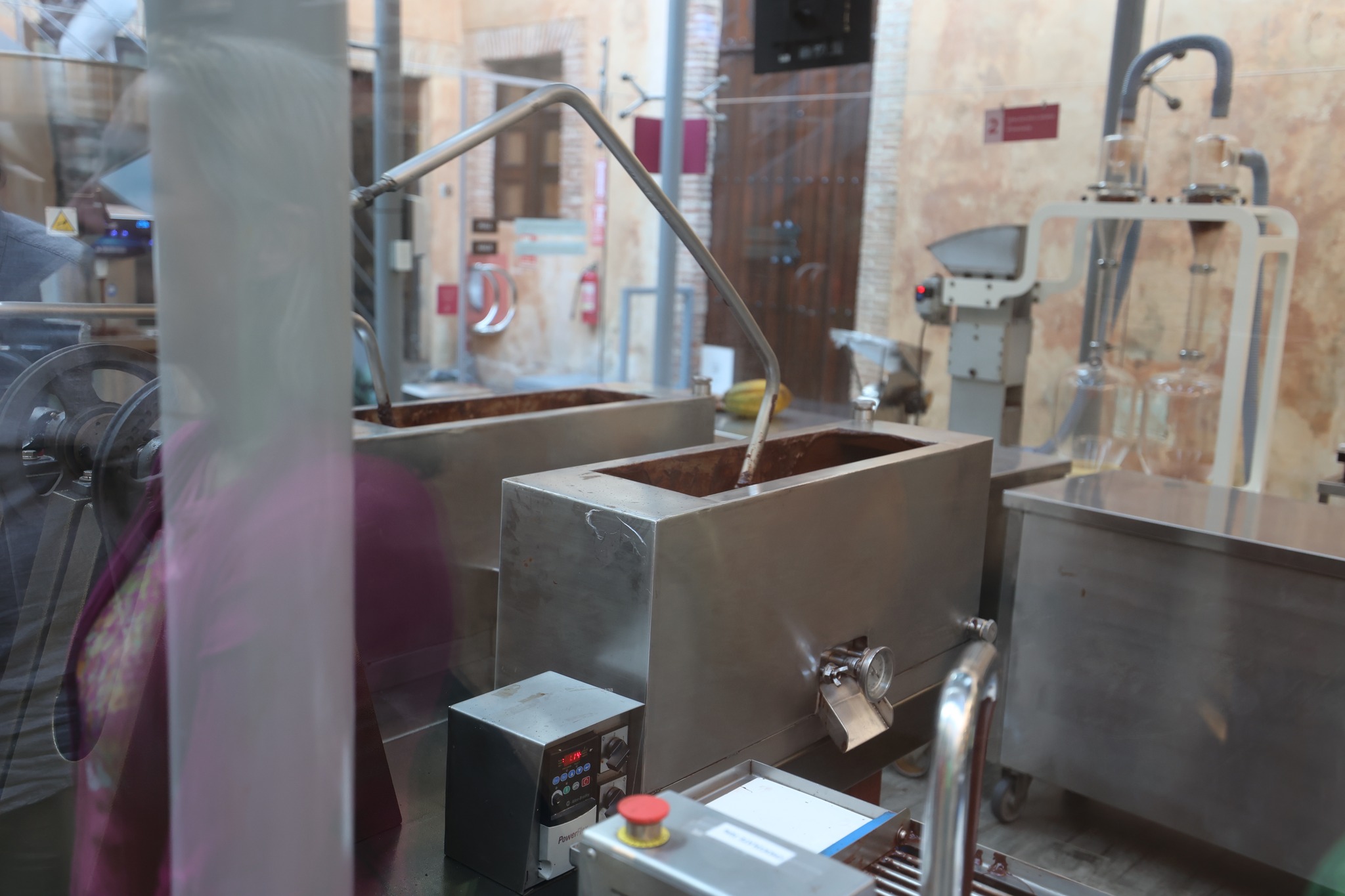 Industrial chocolate processing equipment with stainless steel conching machines behind glass